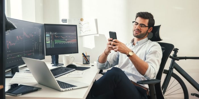 Using modern technologies. Relaxed young trader in formal wear and eyeglasses is using his smartphone while sitting in his office. Forex market. Trade concept. Investment concept